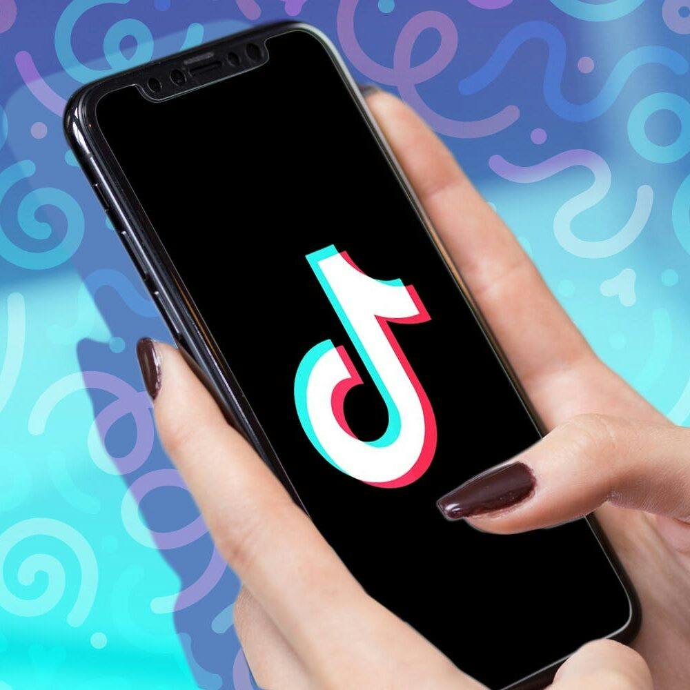 Two hands holding an iPhone showing the TikTok logo in front of a funky, multi-colored backdrop.