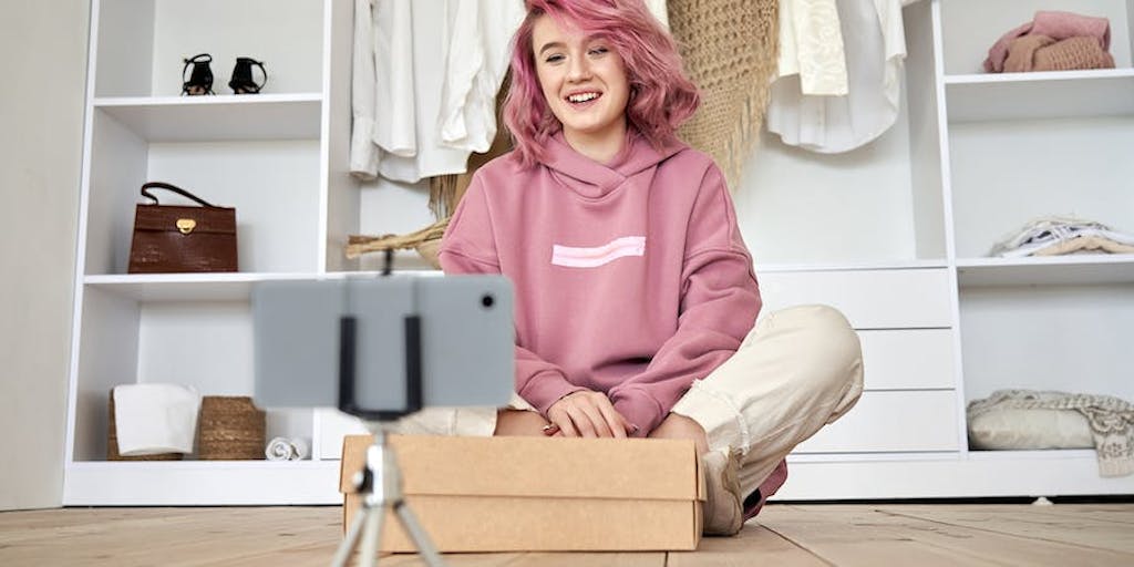 Girl with pink hair sitting cross-legged in front of her closet and filming an unboxing video on her iphone, held up by a tripod