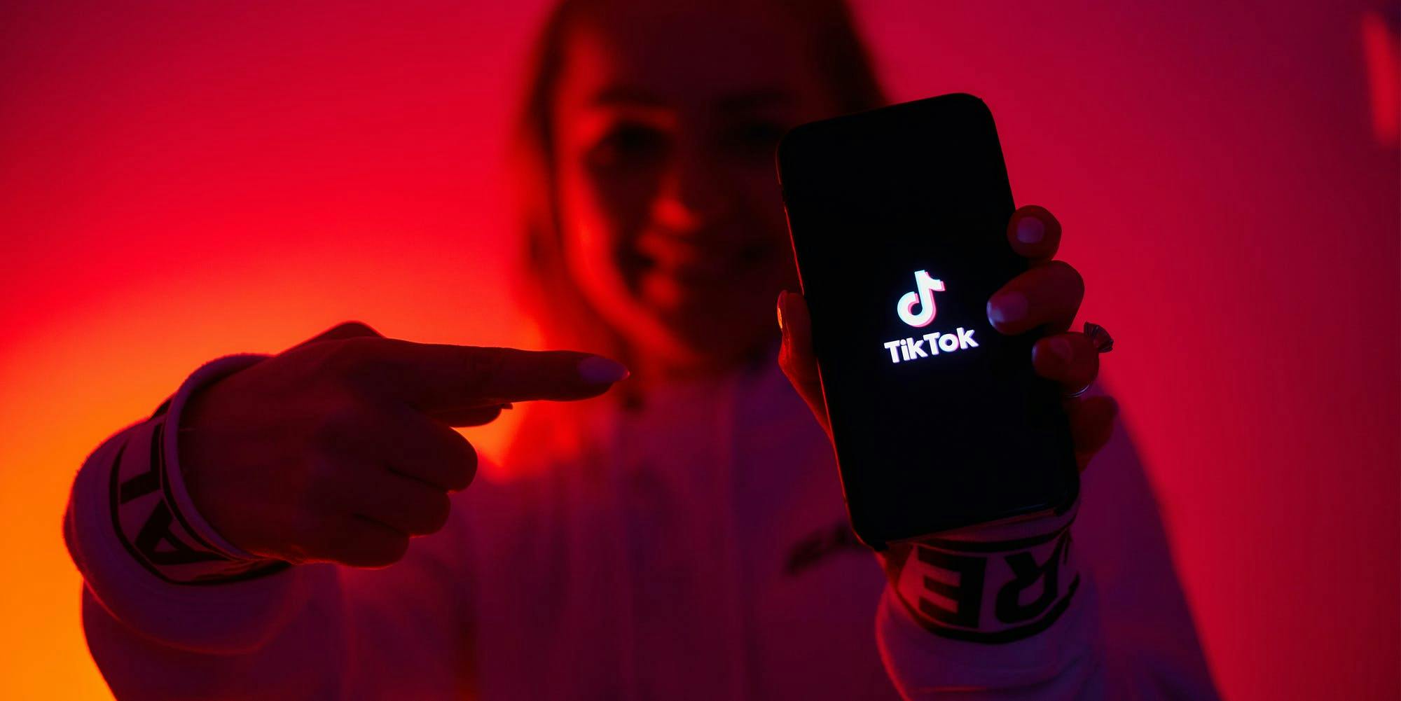 woman holding phone with TikTok on screen while pointing with other hand in front of yellow orange to red radial gradient background