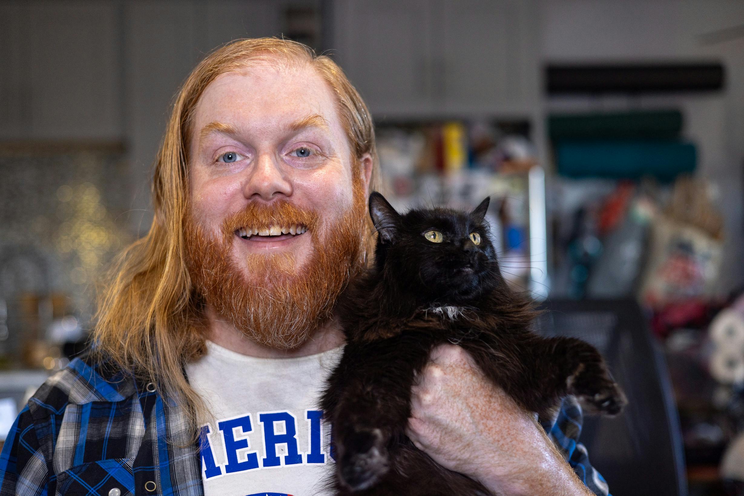 self portrait of a bearded red headed man with long hair and a blue shirt holding a cat
